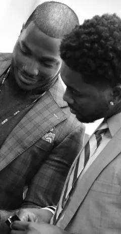 Two men in suits wearing NFLPA lanyards huddled together, looking down at something out of frame.