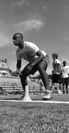 A young football player in a ready stance beside a cone on a stadium field during a drill, with other players visible in the background.
